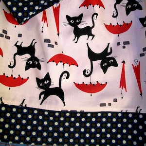 May include: White fabric with a black and white polka dot pattern. The fabric has a red and white umbrella and black cat design. The fabric is folded in half.