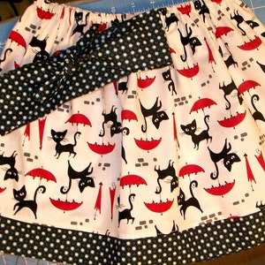 May include: A white skirt with a black and white polka dot headband. The skirt has a black and white print of cats holding umbrellas. The skirt has a black and white polka dot trim.