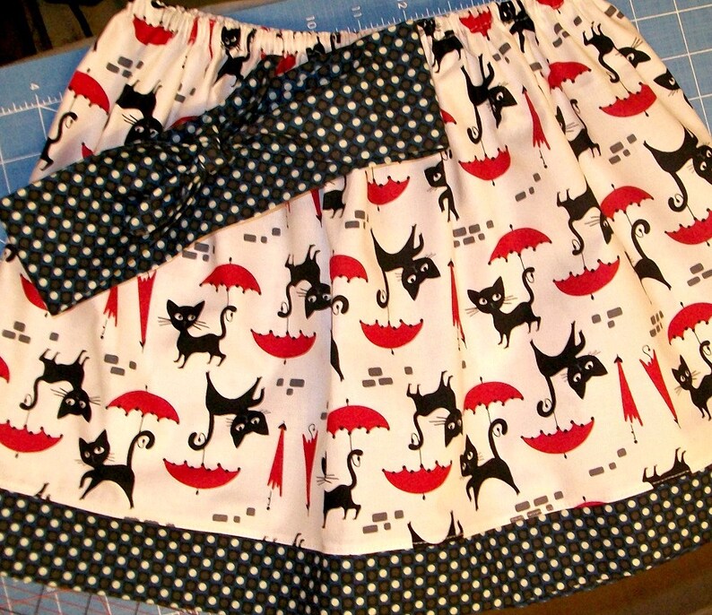 May include: A white skirt with a black and white polka dot headband. The skirt has a black and white print of cats holding umbrellas. The skirt has a black and white polka dot trim.