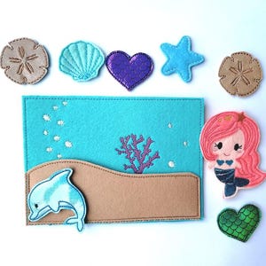 May include: A felt mermaid-themed set featuring a turquoise background with a dolphin, coral, and bubbles. Includes a mermaid, starfish, seashell, heart, and sand dollar appliques. The set is designed with various shades of blue, pink, purple, and brown.