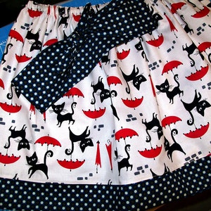 May include: A white skirt with a black and white polka dot pattern. The skirt has a black bow with white polka dots and a red and white umbrella pattern with black cats.