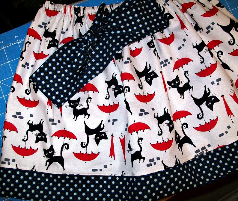 May include: A white skirt with a black and white polka dot pattern. The skirt has a black bow with white polka dots and a red and white umbrella pattern with black cats.