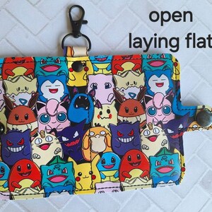 May include: A colorful fabric wallet with a variety of cartoon characters, including Pikachu, Charmander, and Squirtle. The wallet is open and laying flat, showing the inside of the wallet. The wallet has a black metal clip on the top.