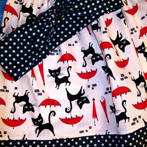 May include: A white skirt with a black and white polka dot bow and black cat and umbrella print. The skirt has a black and white polka dot trim.