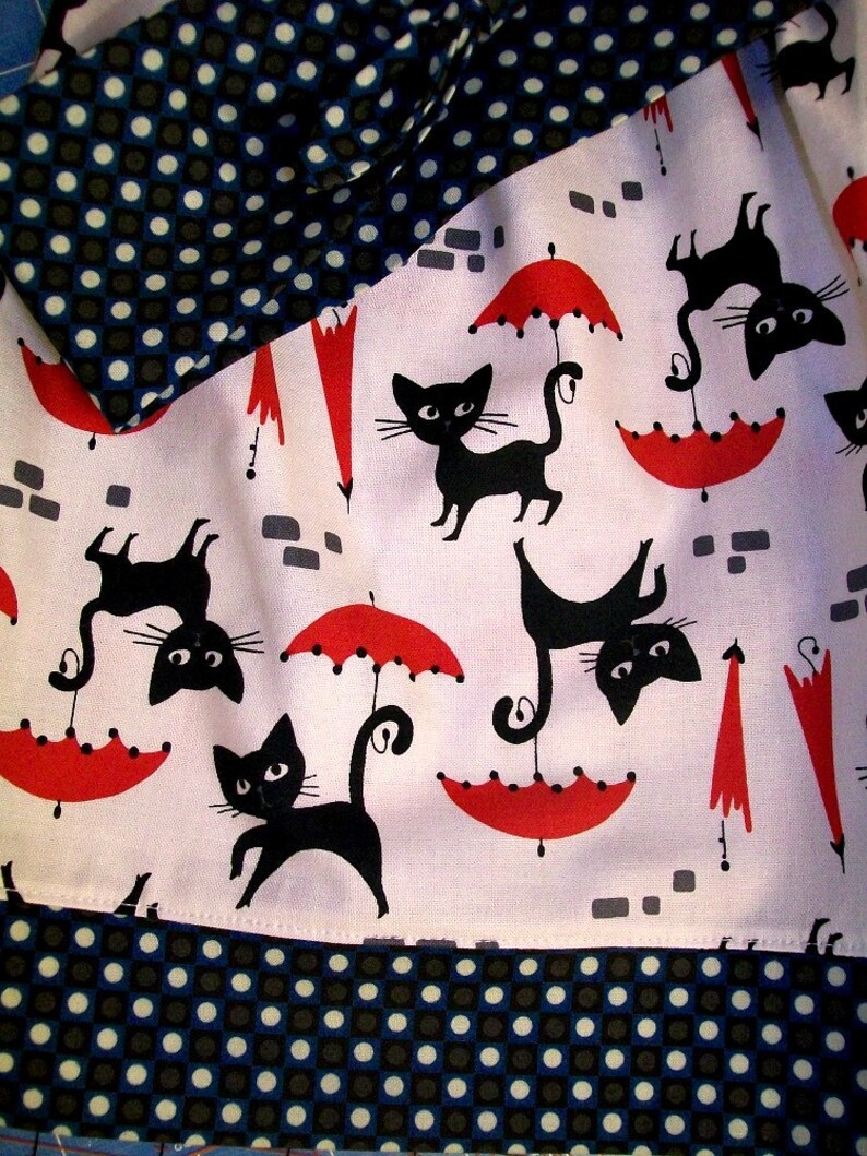 May include: White fabric with a black and white polka dot pattern. The fabric has a red and white umbrella and black cat design. The fabric is folded in half.