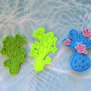 May include: Three felt coral decorations in shades of green and blue with pink accents. The coral shapes are hand-stitched with contrasting thread, and are arranged on a watery background, suggesting an underwater theme.