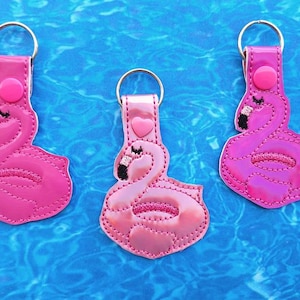 May include: Three pink flamingo pool float keychains with a shiny iridescent finish. Each keychain features a pink flamingo with a black eye and a heart-shaped detail.