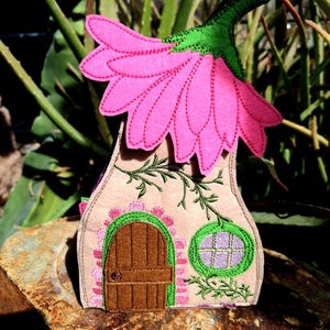 May include: A whimsical felt fairy house with a pink flower roof and green stem. The beige house features a brown door, a round window, and embroidered green vines. The house is decorated with pink and green accents.