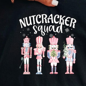 May include: A black t-shirt featuring a white graphic print that reads NUTCRACKER squad with small snowflake accents. Below the text are four illustrated nutcracker figures in shades of pink, white, and blue, each wearing a tall hat and uniform.
