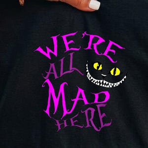 Könnte beinhalten: Ein schwarzes T-Shirt mit dem violetten Schriftzug "WE'RE ALL MAD HERE" in einer stilisierten Schriftart. Rechts neben dem Text befindet sich eine Grafik eines schwarzen Katzengesichts mit leuchtend gelben Augen und einem breiten Grinsen.