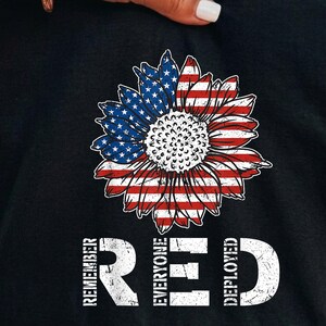 Könnte beinhalten: Ein schwarzes T-Shirt mit einer Sonnenblumen-Grafik, deren Blütenblätter im Design der amerikanischen Flagge mit Sternen und Streifen gehalten sind. Unter der Sonnenblume steht der weiße, abgenutzte Schriftzug REMEMBER EVERYONE DEPLOYED.