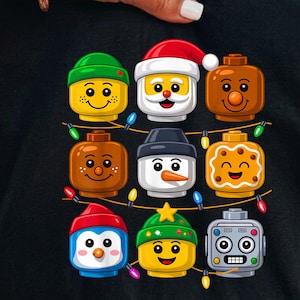May include: A black t-shirt featuring a graphic print of nine stylized toy brick heads arranged in a grid. The characters include Santa, a snowman, a gingerbread person, a robot, and others wearing winter hats, all connected by string lights.