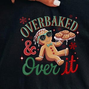 May include: A black t-shirt featuring a graphic of a gingerbread person wearing teal sunglasses and a bow tie. The gingerbread person holds a plate of cookies. The text reads OVERBAKED & Over it, surrounded by red snowflakes and star accents.