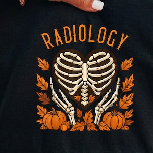 Op de afbeelding: Een zwart t-shirt met een grafische print met het woord RADIOLOGY in oranje letters boven een hartvormige ribbenkast. Twee skeletachtige handen omlijsten het hart, omringd door oranje herfstbladeren en twee oranje pompoenen aan de onderkant.
