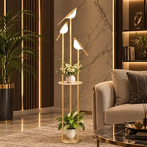 May include: A gold-toned floor lamp featuring three vertical poles of varying heights, each topped with a white bird-shaped light fixture. The lamp includes a circular shelf holding a small white vase with flowers and a base holding a potted plant.