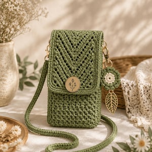 May include: A sage green crochet crossbody bag featuring a chevron pattern on the flap, a circular gold button with a botanical line drawing, and a matching long strap. A small crocheted flower charm and a gold metal leaf pendant are attached.