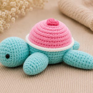 May include: A crocheted turtle toy featuring a light blue head and flippers, a bright pink shell with a small heart-shaped accent on top, and a white border separating the shell from the body. The turtle has two small black bead eyes.
