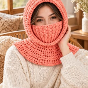 May include: A coral-colored crochet balaclava featuring a thick, ribbed texture that covers the head, neck, and lower face. The headpiece has a wide, rounded hood opening and a fitted, vertical-ribbed section that extends over the nose and mouth.