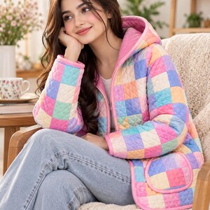 Peut inclure: Une veste matelass&eacute;e &agrave; capuche avec un motif &agrave; carreaux multicolores dans des tons de rose, bleu, jaune et violet. La veste pr&eacute;sente une fermeture &eacute;clair frontale, des manches longues et deux poches avant avec un liser&eacute; rose, port&eacute;e sur un jean bleu.