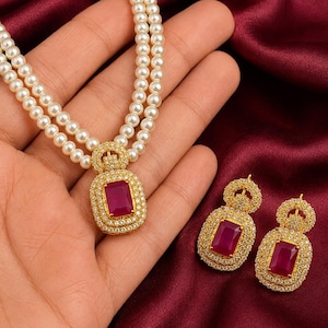 Op de afbeelding: Een sieradenset bestaande uit een dubbelstrengs ketting en bijpassende oorbellen. De ketting is voorzien van witte parels en een goudkleurige hanger met een rechthoekige rode steen omringd door kleine heldere steentjes.