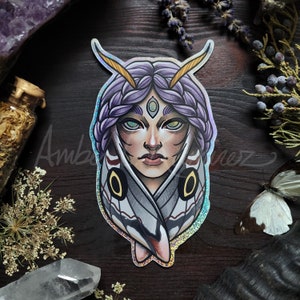 May include: A holographic sticker featuring a woman with purple hair and a third eye, surrounded by the wings of a moth. The wings are white with black and red markings.