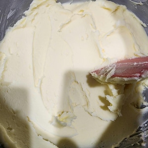 May include: A stainless steel mixing bowl containing a smooth, pale yellow creamed butter mixture. A red silicone spatula is positioned on the right side, pressing into the thick, textured substance, showing clear streaks and ridges from the mixing process.