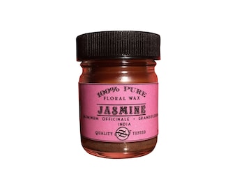 May include: A small glass jar of jasmine floral wax with a black lid. The jar has a pink label with the words "100% Pure Floral Wax Jasmine" in white and black text. The bottom of the jar contains a brown wax.