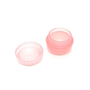 May include: A small, open, pink plastic jar with a matching lid. The jar is round with a slightly bulbous shape and a threaded rim. The lid is flat and circular. The jar and lid are set against a white background.
