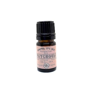 May include: A small amber glass bottle of 100% pure patchouli essential oil with a black ribbed cap. The label is light pink with black text reading 100% PURE ESSENTIAL OIL PATCHOULI IRON DISTILLED POGOSTEMON CABLIN INDIA QUALITY TESTED.