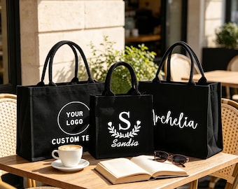 Könnte beinhalten: Drei schwarze Tragetaschen aus Segeltuch mit weißen Aufdrucken. Die linke Tasche zeigt ein kreisförmiges Logo, die mittlere Tasche einen Buchstaben S mit einem Blumenkranz und dem Namen Sandia, die rechte den Namen Archelia.