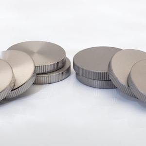May include: A collection of circular, metallic discs with a brushed finish and a textured, ribbed edge. The discs are arranged in two small stacks and several individual pieces, all featuring a uniform silver-grey color and a flat top surface.