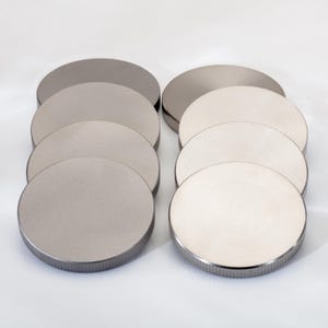 May include: Eight circular metal discs arranged in two vertical columns. Each disc features a smooth, flat top surface and a textured, ribbed edge. The discs on the left have a matte grey finish, while the discs on the right have a polished, reflective silver finish.