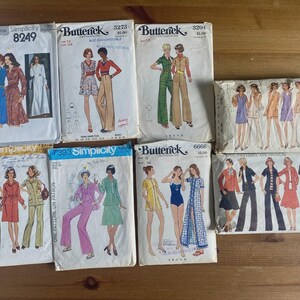 May include: A collection of eight vintage sewing pattern envelopes from Simplicity and Butterick brands, featuring various illustrations of clothing styles including dresses, jumpsuits, and suits with printed model numbers and brand logos.