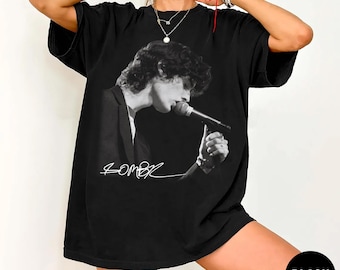 May include: A black short-sleeved t-shirt featuring a monochromatic graphic print of a person with curly hair singing into a microphone while wearing a suit jacket. Below the figure, a white handwritten-style signature is printed on the fabric.