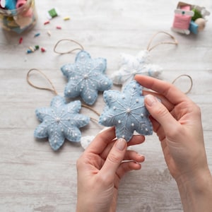 May include: A collection of handmade felt snowflake ornaments in shades of light blue and white. Each snowflake features intricate white embroidered details and small white beads. The ornaments include thin twine loops for hanging purposes.