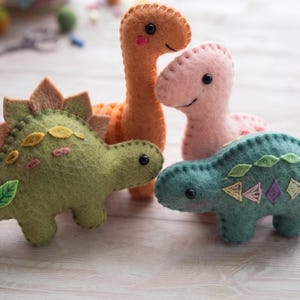 May include: Four felt dinosaur plush toys in various colours including olive green, orange, light pink, and teal. Each dinosaur features stitched details, small black bead eyes, and embroidered patterns along their backs and sides.