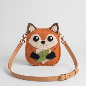 May include: A felt crossbody bag shaped like a fox face with orange and cream coloring. The fox features black eyes, a small nose, and black paws holding a green felt leaf. It includes a tan leather adjustable strap with metal hardware.