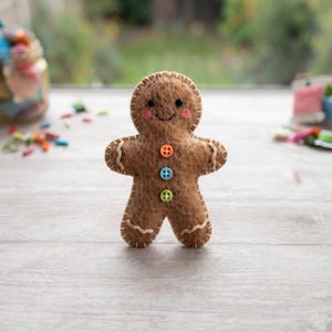 May include: A brown felt gingerbread person doll with a fuzzy texture, featuring a stitched cream-colored border, two black bead eyes, a stitched smile, pink cheeks, and three small buttons in orange, blue, and green on the torso.