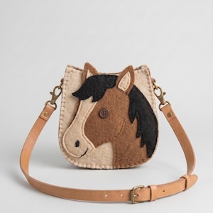 May include: A felt crossbody bag featuring a brown and cream horse head applique with a black mane and a brown button eye. The bag has a light tan leather strap with brass-toned metal hardware, including swivel clips and an adjustable buckle.