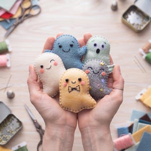Könnte beinhalten: Fünf kleine, handgefertigte Filzgeister in verschiedenen Farben wie Creme, Blau, Hellgrün, Gelb und Grau. Jeder Geist hat gestickte Gesichtsausdrücke, rosa Wangen und einzigartige Details wie eine Fliege oder bunte Sternenverzierungen.