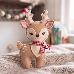 Può includere: Un peluche in feltro color marrone chiaro a forma di cerbiatto con corna color crema, macchie bianche sul dorso e una sciarpa a quadretti rossi e bianchi annodata al collo con un piccolo bottone in legno e un campanellino in metallo.