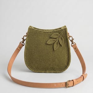Puede incluir: Un bolso bandolera de fieltro verde oliva con base redondeada y una aplicación en forma de hoja en la parte delantera. El bolso cuenta con una correa ajustable de cuero color canela sujeta con mosquetones de metal color latón.