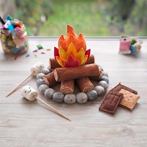 May include: A felt campfire set featuring brown logs, a layered orange, yellow, and red flame, and a ring of grey stones. Two white felt marshmallows with embroidered smiling faces on wooden skewers and two brown and tan felt s'mores are included.