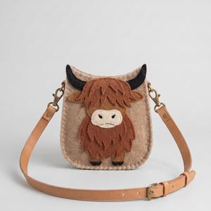 Peut inclure: Un sac bandoulière en feutrine beige orné d'une application de vache Highland marron avec des cornes noires. Le sac est doté d'une bandoulière en cuir beige avec des ferrures en métal doré, incluant des mousquetons et une boucle réglable.