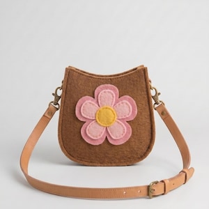 May include: A brown felt saddle-shaped crossbody bag featuring a large, layered pink and yellow felt flower appliqué on the front. The bag includes a tan leather adjustable shoulder strap attached with brass-toned metal lobster claw clasps.