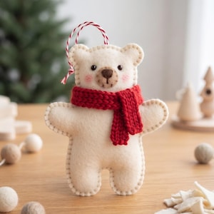 May include: A cream-colored felt bear ornament featuring a hand-stitched edge, a dark brown embroidered nose and mouth, and pink blush on the cheeks. The bear wears a bright red knitted scarf and has a red and white twisted string loop for hanging.