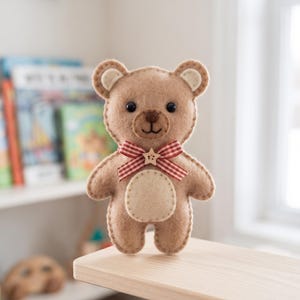 Puede incluir: Un oso de peluche de fieltro color canela con un parche en el vientre de color crema, ojos redondos oscuros, nariz y boca bordadas, y un lazo de cinta de cuadros rojos y blancos en el cuello adornado con un pequeño botón de madera en forma de estrella.