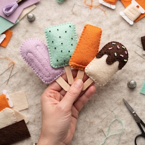 May include: Four felt ice cream treats on wooden sticks, including a lavender popsicle, a mint green rectangular bar with black seed beads, an orange popsicle, and a cream-colored bar with brown felt topping and colorful seed bead sprinkles.