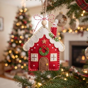 May include: A red felt house ornament featuring a white roof, a brown door with a yellow knob, two white windows, a green wreath, and a chimney with white smoke. The ornament is suspended by a jute string with a red and white twine bow.