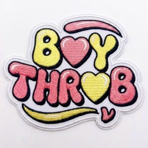 May include: An embroidered patch featuring the text BOY THROB in a bubble font. The letters are yellow and pink with thick black outlines. A pink heart replaces the O in BOY and a yellow heart replaces the O in THROB. Pink and yellow swooshes accent the design.